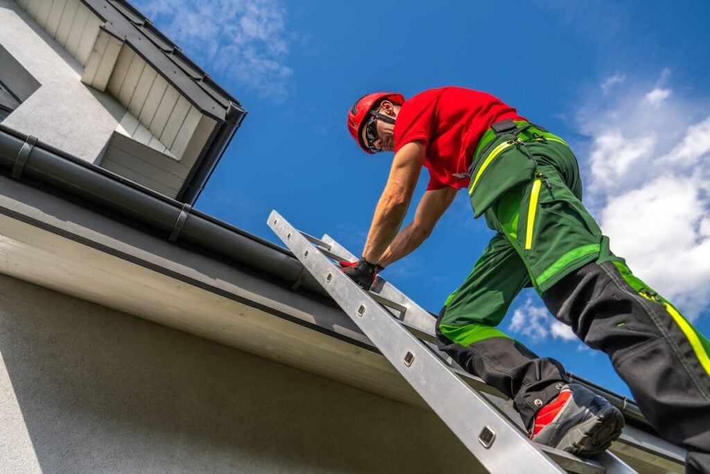 A maintenance worker in safety gear ascends a ladder to inspect and repair a roof gutter while the sun shines brightly overhead. vinyl siding cost
