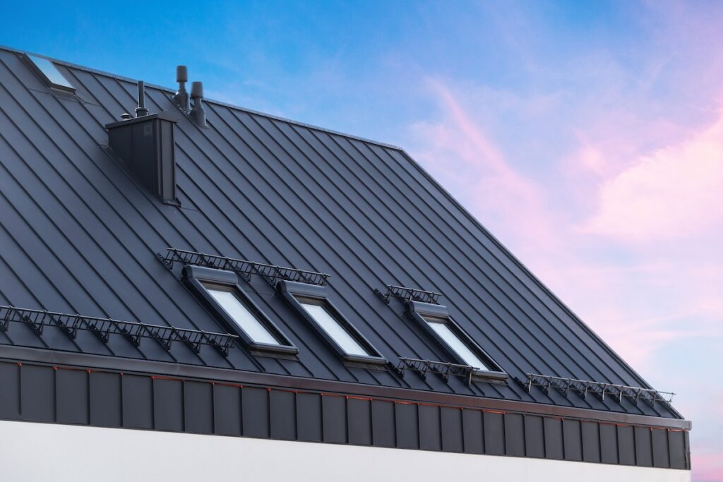 standing seam metal roof House with metal galvanized coating, chimney pipe, mansard windows and snow holding cornice on roof slope against sunset sky. Element of modern residential building with zinc rooftop sheets